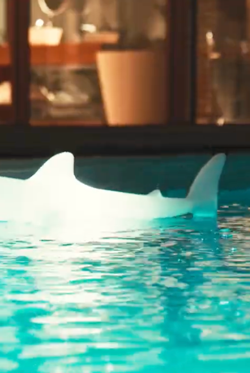 Goodnight Light Bobb the pool shark lamp glowing in a turquoise swimming pool, evening lights on in the villa windows behind