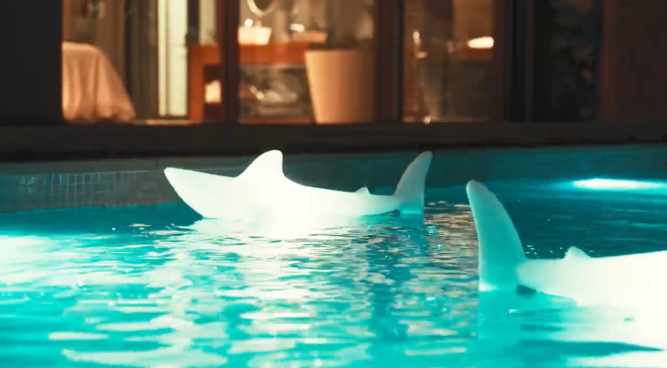 Goodnight Light Bobb the pool shark lamp glowing in a turquoise swimming pool, evening lights on in the villa windows behind